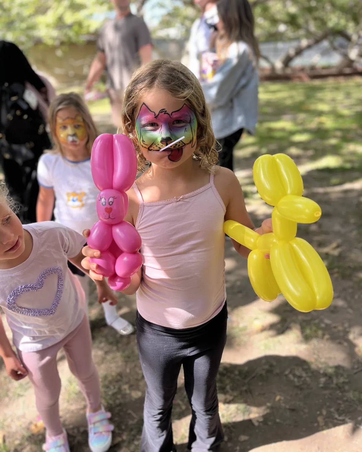 Child holding pink and yellow balloon animals at an outdoor Happy Faces LA party